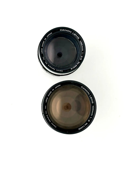 Two black canon camera lenses, one 50mm f/1.4 and the other 100mm f/2, with silver lettering on their rings.