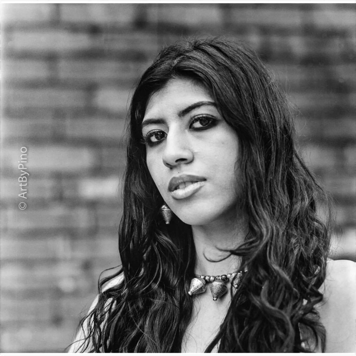 A black-and-white portrait of a woman with long, wavy hair wearing an ornate metallic necklace.