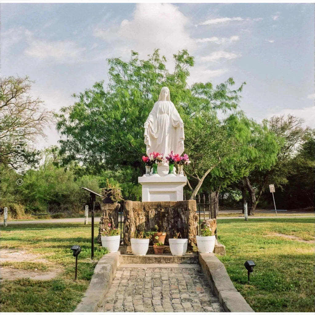 White marble statue of the virgin mary standing on a stone pedestal, adorned with pink flowers in green vases.