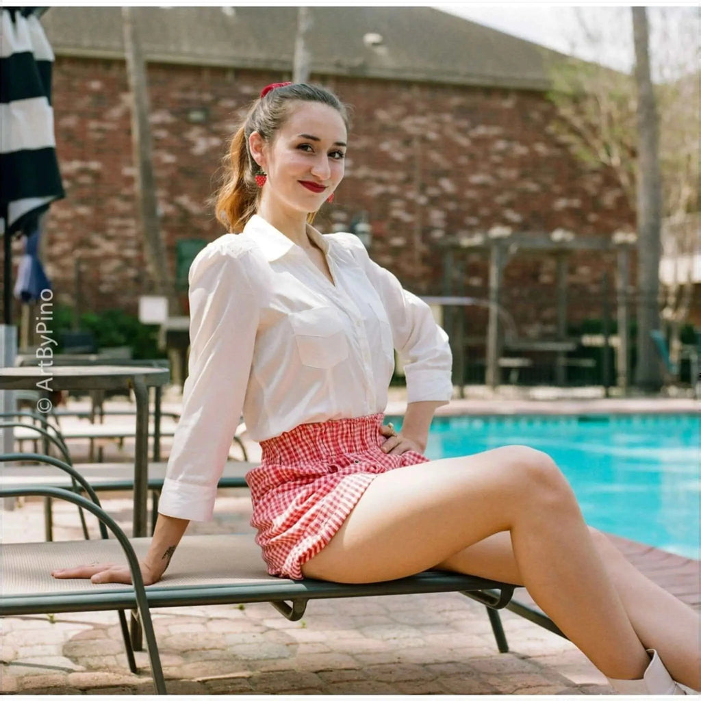A woman wearing a white button-up shirt and red-and-white gingham shorts sits on a lounge chair by the pool.