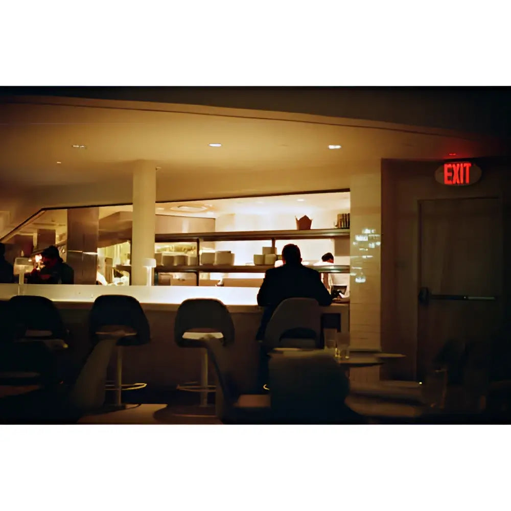 The image shows a dimly lit restaurant interior with patrons seated at bar stools, and an illuminated red ’exit’ sign above a door on the right.